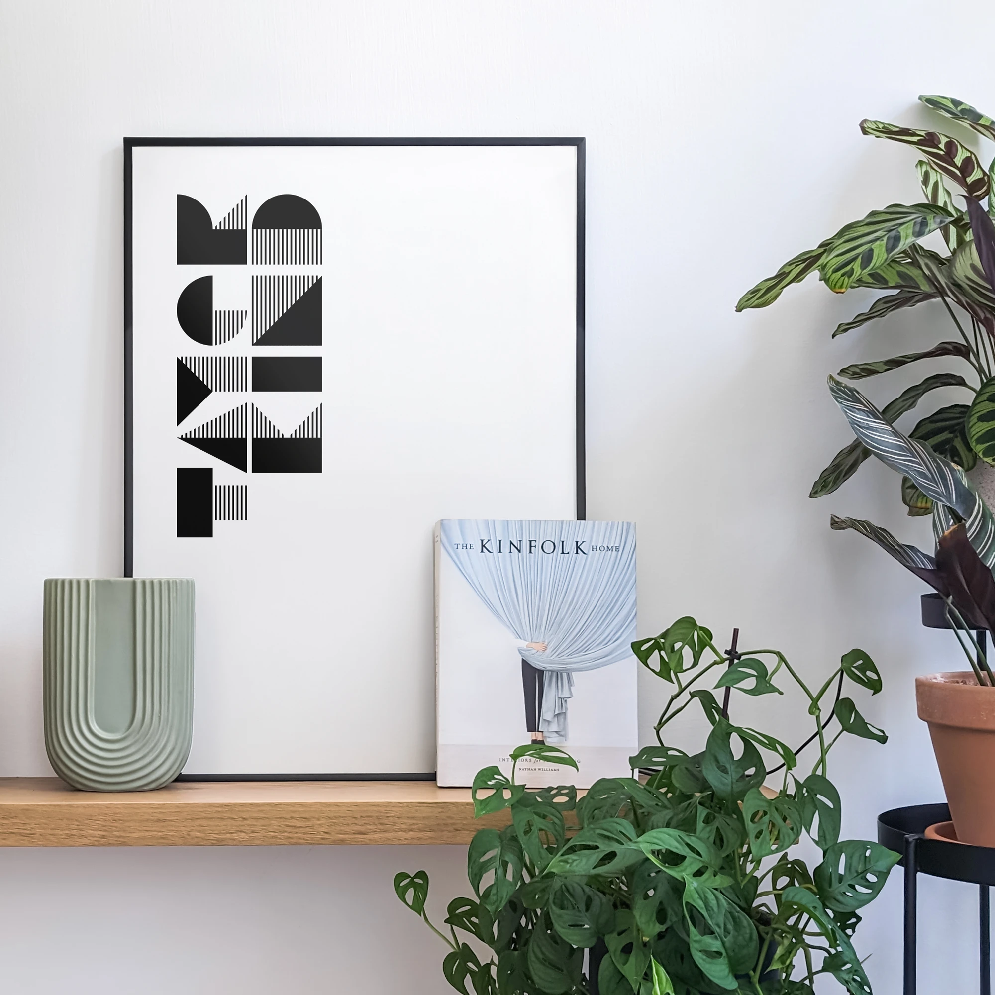 פוסטר גרפי של המילה Tayer Kind ביידיש, מעוצב בסגנון באוהאוס, נשען על מדף עץ ולצידו פריטי עיצוב ועציצים