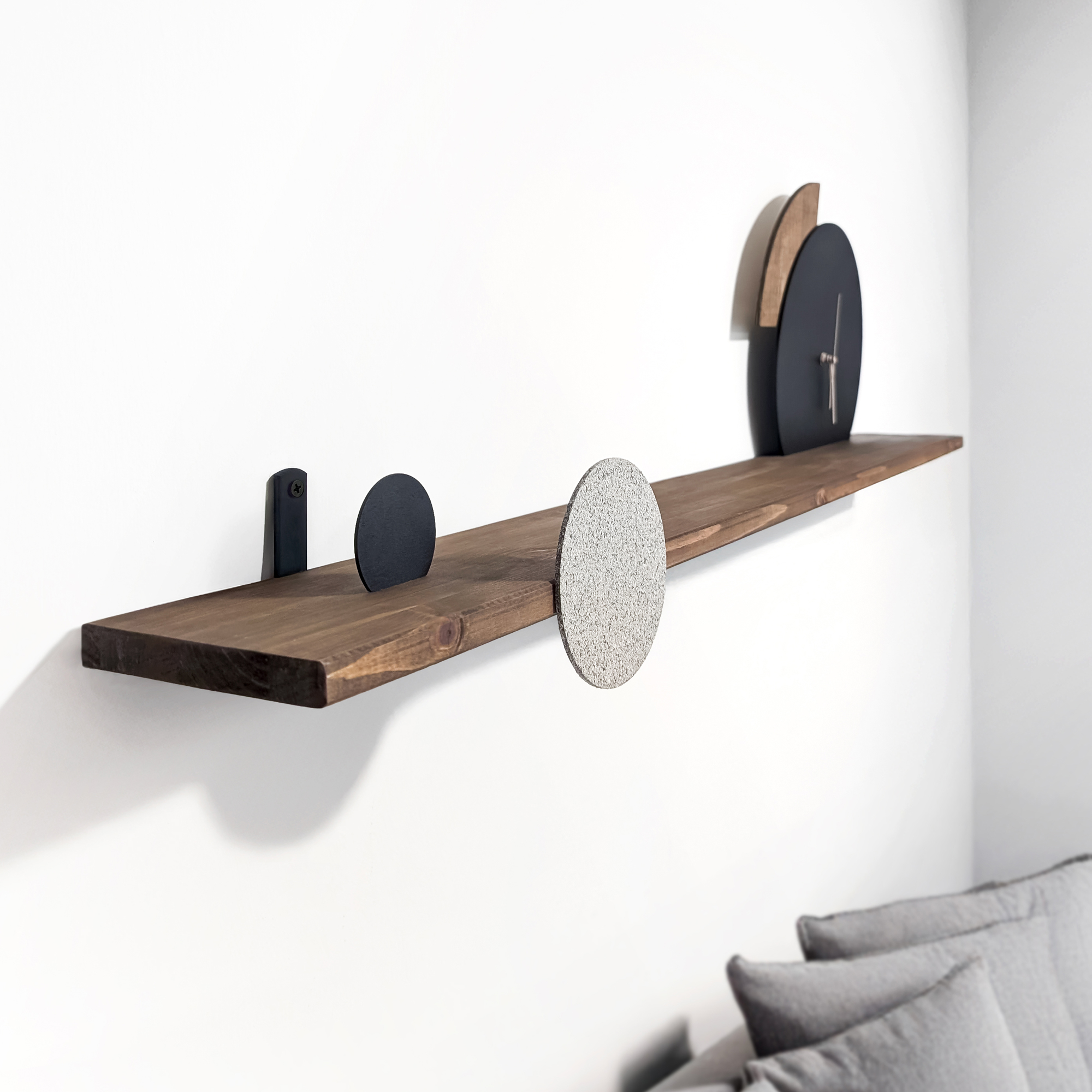 מדף עץ משולב שעון גיאומטרי מעוצב אישית בגווני שחור, טקסטורת אבן ועץ עם מחוגים בגוון פודרה, תלוי על קיר מעל ספה אפורה