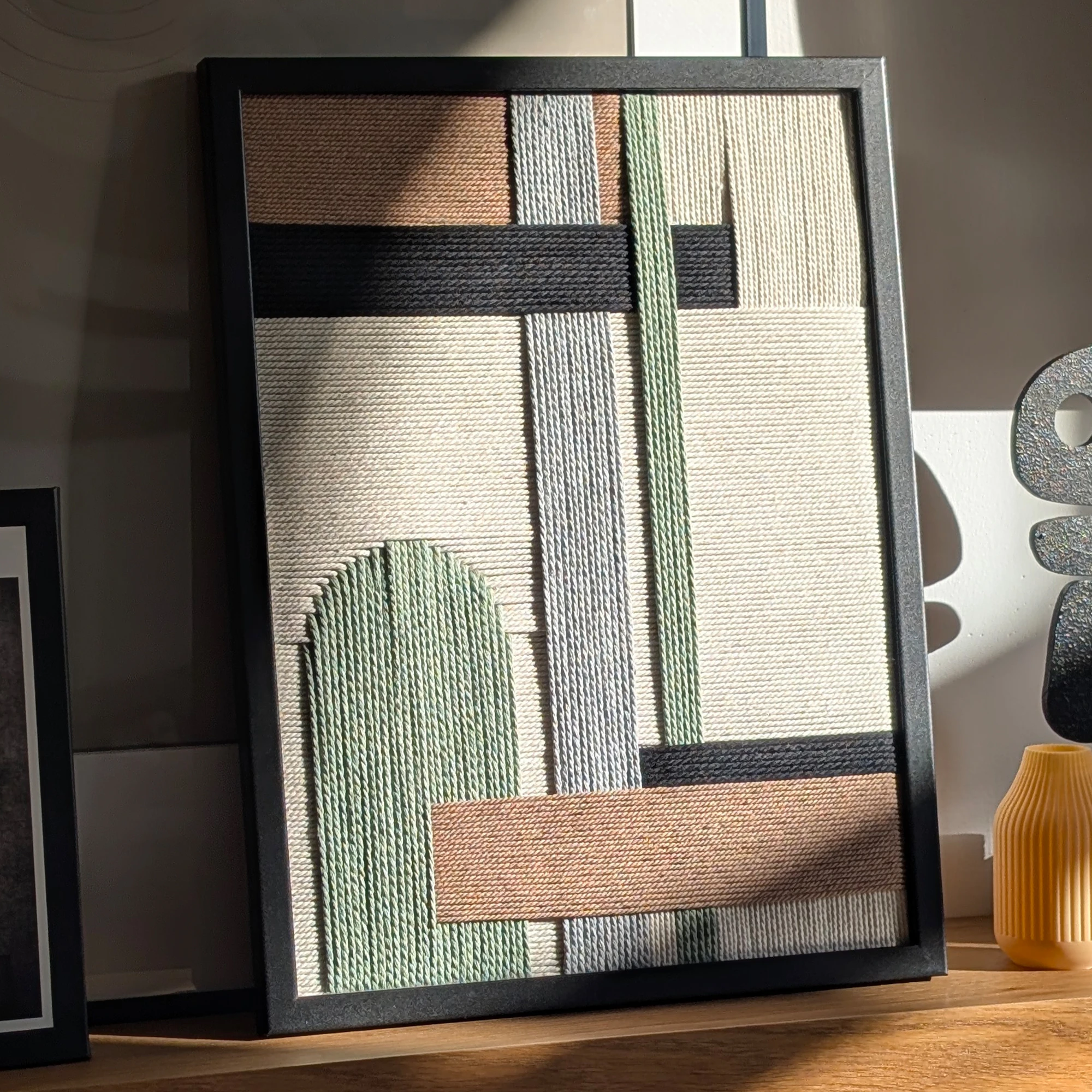 תמונה ממוסגרת בשחור של אריגת חוטי כותנה בגווני שחור, אפור, חום, ירוק מרווה וגוון טבעי, מונחת על מדף מואר בקרן שמש