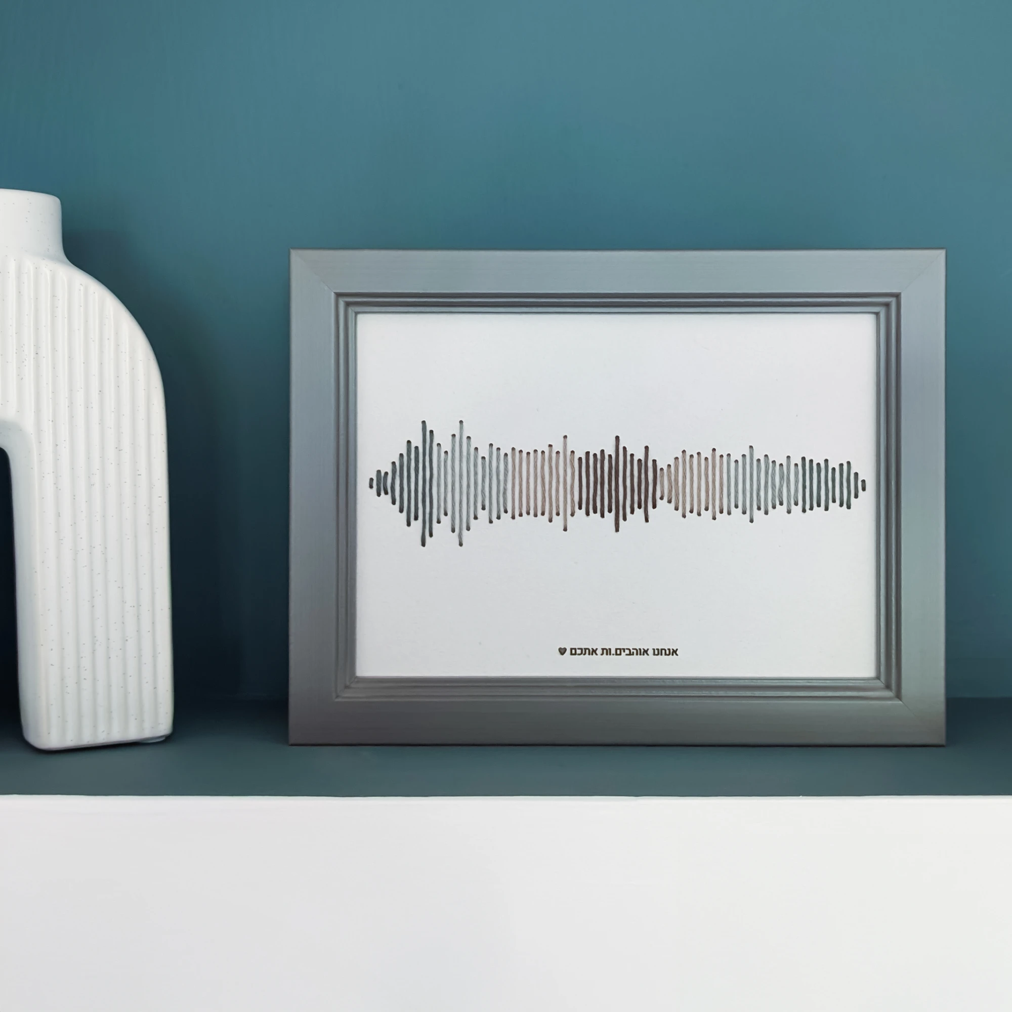 רקמת כותנה של פסקול הקלטה אישית בגווני ירוק ואפור, עם מסגרת אפורה וחריטת טקסט בתחתית, מונחת על מדף ירוק
