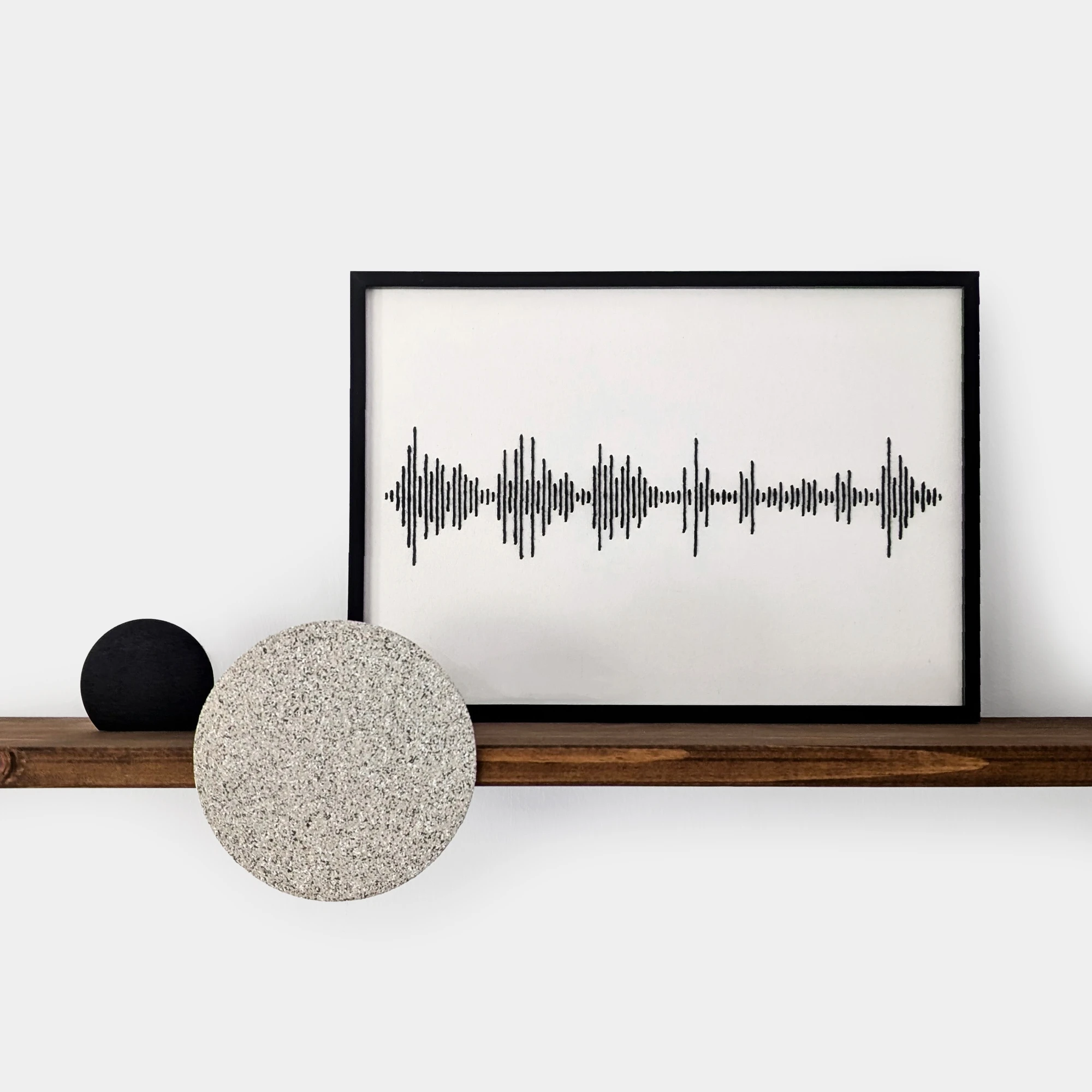 רקמת כותנה שחורה של פסקול הקלטה אישית ממוסגרת במסגרת שחורה, שעונה על מדף עץ עם עיגולים דקורטיביים