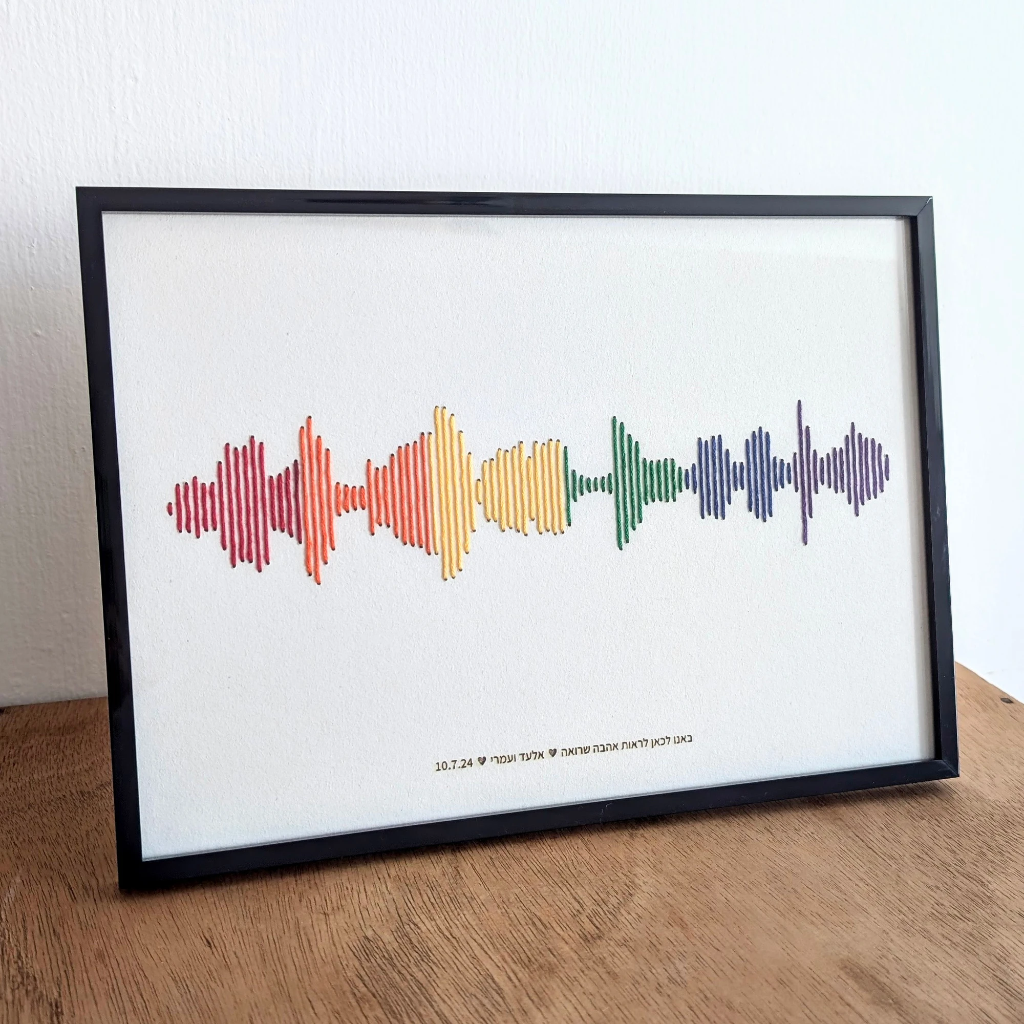 רקמת כותנה של פסקול הקלטה אישית בצבעי גאווה, עם מסגרת שחורה וחריטת טקסט בתחתית, מונחת על שידת עץ