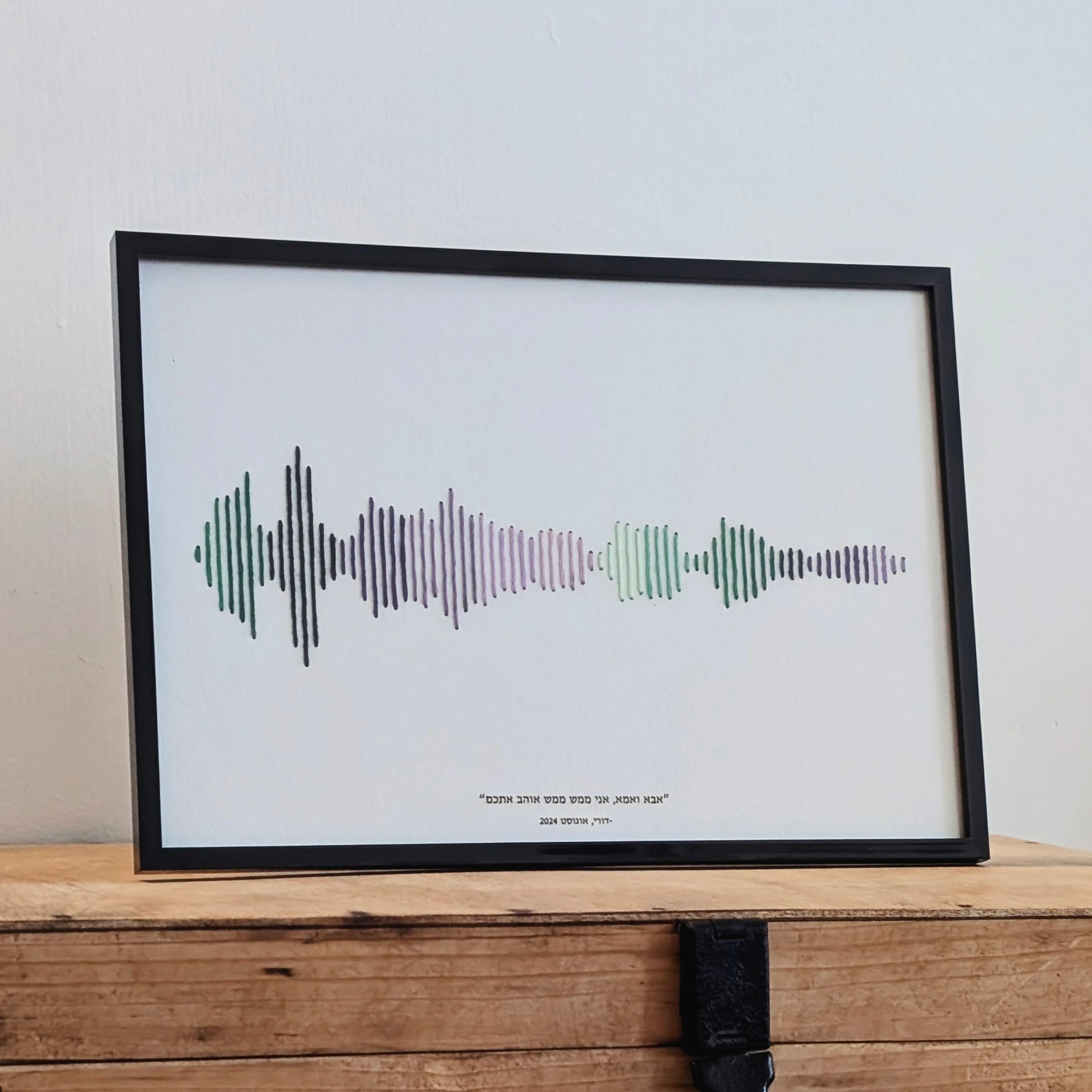 רקמת כותנה של פסקול הקלטה אישית בגווני ירוק וסגול, עם מסגרת שחורה וחריטת טקסט בתחתית, מונחת על שידת עץ